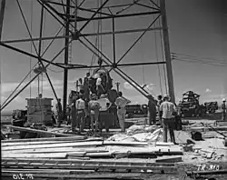 Hommes rassemblés autout d'une large structure en forme de derrick, un large objet rond est en train d'être soulevé.