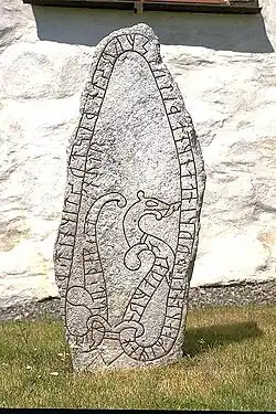 Photo d'une stèle de pierre de couleur gris clair aux bords irréguliers. Elle est dressée sur un sol herbeux, devant un mur blanchi à la chaux. Un ruban est gravé sur tout le pourtour de la pierre et s'incurve à l'intérieur pour figurer un animal. Des runes sont gravées à l'intérieur du ruban.