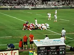 Sur la pelouse d'un stade, deux équipes de joueurs de rugby disputent une mêlée. En arrière-plan, des spectateurs sont assis dans les tribunes.
