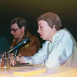 Le Guin, assise à une table de conférence, parle dans un micro, pendant qu'à sa gauche l'écrivain Harlan Ellison l'écoute