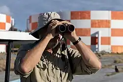 Un homme barbu et portant un bob regarde dans des jumelles sur une plage à proximité de grands réservoirs d'eau