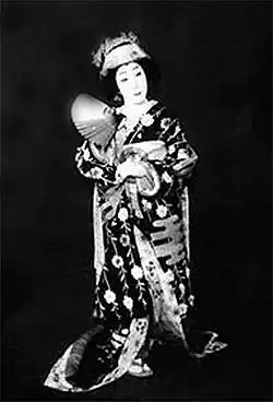 Portrait en noir et blanc d’un homme qui porte un kimono sombre avec des motifs floraux et un col blanc. Il a les cheveux noirs coiffés en chignon et le visage maquillé de blanc. Il regarde vers la droite avec une expression sérieuse.