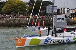 Voilier blanc descendant au moteur un chenal. Foule massée sur le quai.