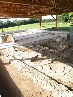 Au premier plan, les vestiges archéologiques principalement des murs et en arrière plan le toit du préau en bois
