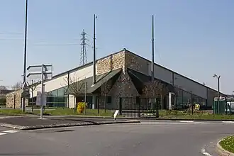 Vue d'un bâtiment moderne partiellement en meulière et à toiture à facettes ; deux mâts près des entrées.