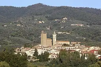 Photographie en couleurs représentant un village sur le flanc d'une colline. Les maisons ont une façade beige et des toits rouge ou rouille. Une église et un clocher surplombent le village.