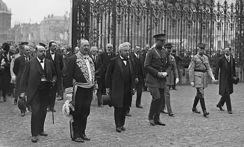 Photo en noir et blanc d’un groupe d’hommes (civils et militaires) pénétrant dans une cour entourée d'une grille