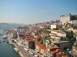 Une ville côtière très ensoleillée sur la droite, construite à flanc de colline. Les nombreuses maisons blanches, jaunes ou rouges ont toutes des toits de tuiles oranges. L'eau d'un estuaire et quelques petits bateaux sont visibles au pied de la colline sur la gauche