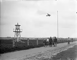 Photo noir et blanc d'un biplan qui décolle et de personnes marchant en lisière de la base.