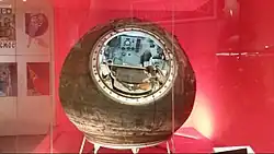 Capsule spatiale exposée derrière une vitrine, avec l'intérieur de visible et contenant de nombreux gadgets électroniques et un siège