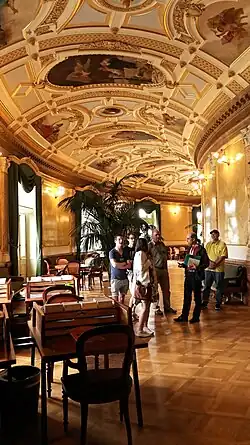 Photo d'une salle courbe avec dorures, sculptures, tableaux sur le plafond, de même que des tables de travail et chaises en bois.