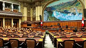 Photographie d'une grande salle munie de pupitres et de chaises, au fond une tribune et sur la droite en haut une grande peinture représentant un paysage