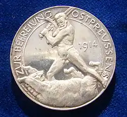 Le revers d'un médaillon d'argent allemand de la Première Guerre mondiale célébrant la libération de la Prusse orientale en 1914 par Paul von Beneckendorff und von Hindenburg, se référant à la bataille de Tannenberg. C'est une représentation du général Hindenburg nu combattant un ours russe avec son épée.