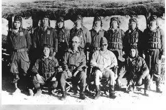Photo noir et blanc d'un groupe d'hommes, pour la plupart en tenues de pilotes, debout, assis ou accroupis