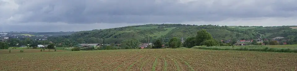 Panorama de la commune, dans la vallée au centre (église, habitat, constructions liées à l'activité humaine. Le premier plan est formé par un champ ; l'arrière plan par les coteaux calcaires, composé de zones forestières et ouvertes, et d'un ciel nuageux