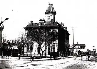 Photo en noir et blanc d'un bâtiment avec clocher en bordure de route. Sont présents deux chevaux tirant des chariots.