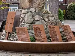 Photographie en couleur montrant un amas de pierres entouré de plusieurs plaques de métal rouillé, gravées avec des noms
