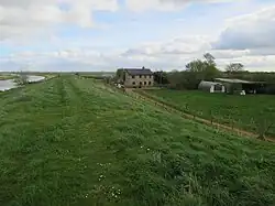 Vue panoramique sur ferme rurale : champ vert, bâtiments en pierre et agricoles, arbres, ciel nuageux.