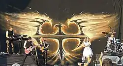 Photo en couleur d'un groupe de musique sur scène, à l'arrière-plan un logo argenté sous forme d'un oiseau de feu