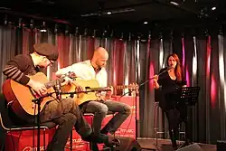 Photo en couleur d'un groupe jouant de la musique dans une pièce, deux guitaristes sont assis à gauche, une chanteuse aux cheveux bruns est debout derrière un pupitre à droite