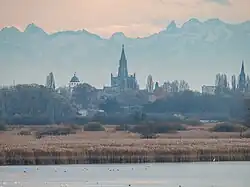 Photo prise par temps nuageux montrant au premier plan un cours d'eau puis de la végétation, joncs et arbres, derrière un village et à l'arrière-plan une chaîne de montagne