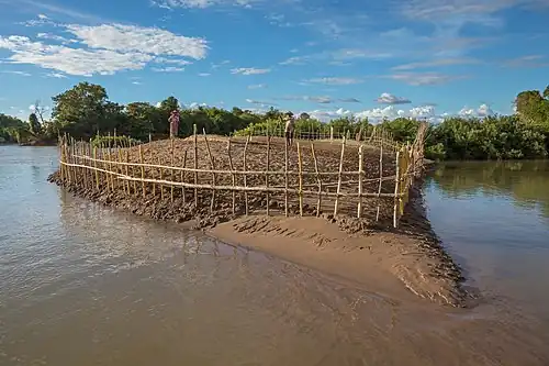 Clôture en bois sur une berge du Mékong, autour d'une petite île cultivée près de Don Loppadi, avec un couple travaillant, à l'heure dorée. Cette barrière est une protection contre les intrus qui voudraient cultiver le même terrain. Cette petite terre n'appartient certainement à personne officiellement, ou bien le propriétaire officiel ne s'en soucie pas. Du fait que cette petite île est totalement immergée pendant la mousson, les premiers locaux qui décident de s'établir là après la décrue du Mékong doivent donner un signal clair au voisinage que le domaine n'est plus disponible.