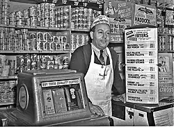 Photographie en noir et blanc d’un épicier souriant, cravaté, portant un tablier blanc à bretelle unique passant derrière le cou, coiffé d’un calot publicitaire ; l’homme s’appuie à une machine distributrice de café et à un comptoir surmonté de panneaux publicitaire ou annonçant les prix de marchandises. Derrière lui, l’étagère qui court tout le long du mur est remplie d’une multitude de boîtes de conserve, parfois cachées par des calicots publicitaires supplémentaires, donnant l’impression qu’il n’y a plus 1&nbsp;cm2 de disponible.