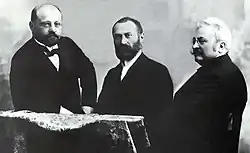 Noir et blanc, les trois sont habillés en costume noir avec chemise blanche et nœuds papillons noirs. Les deux de gauche ont une barbe et sont bruns, celui de droite à une moustache et des cheveux blancs.