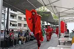Parade rouge comprenant des échassiers, des danseurs et des musiciens percussionnistes.