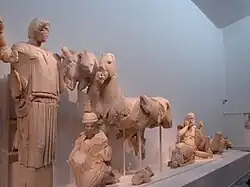 Fronton est, partie droite. Hippodamie soulève un pan de son péplos, geste rituel de la jeune mariée ; une jeune femme, agenouillée, s'occupe des chevaux ; un devin (Clytios, Iamos ou Amythaon), le visage exprimant l'angoisse, car il a prévu l'issue de la course ; la figure suivante est celle d'un enfant jouant avec son orteil ; enfin, la personnification du fleuve Alphée (ou Kladéos) fait l'angle du fronton.