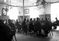 Photographie noir/blanc d’hommes, attablés dans une salle; trois grandes fenêtres offrent une forte lumière en arrière-plan