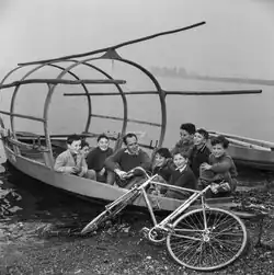 Photographie en noir et blanc de Koblet assis dans une barque au milieu d'enfants, sur les rives d'un lac, son vélo posé devant.