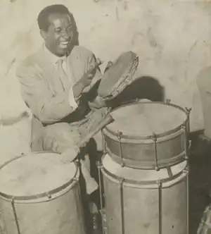 Photographie en noir et blanc d'un homme assis, vêtu d'un complet. Il a un mur dans le dos. Devant lui sont posés trois instruments de percussions. Il tient dans sa main gauche un autre instrument de percussion, plus petit que les autres, peut-être un tamborim. Sa main droite est levée, prête à frapper l'instrument. L'homme a la bouche ouverte.